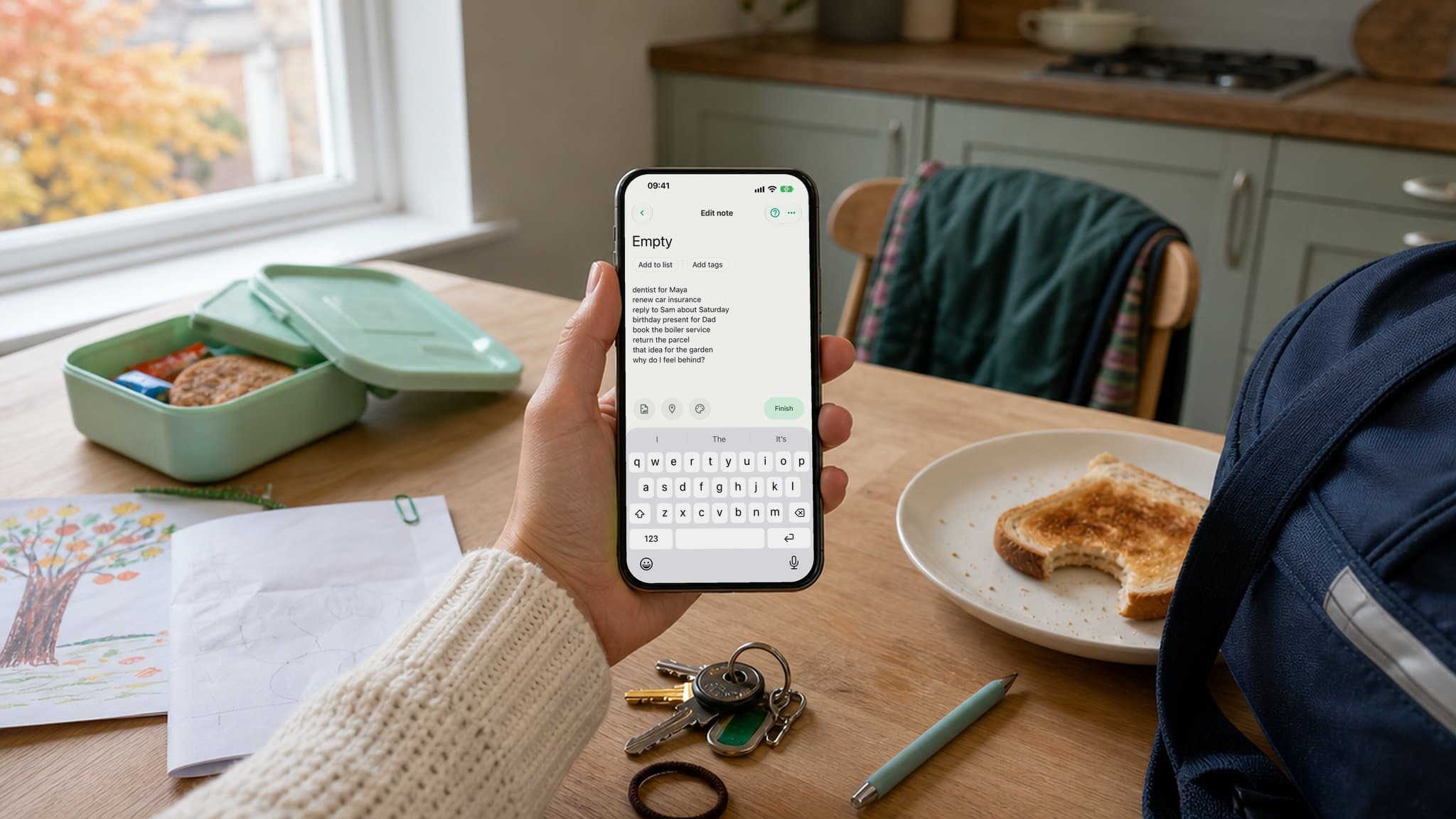 Une main tient un téléphone affichant une note intitulée Empty dans Do Everything, au-dessus d’une table de cuisine un matin d’école, avec une boîte à goûter, une tartine, des clés et un dessin d’enfant